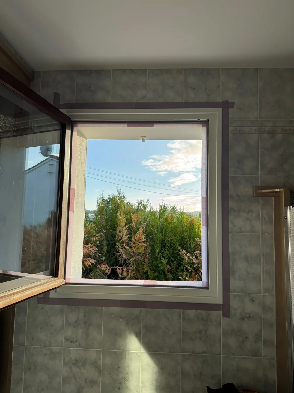 Encadrement de fenêtre de salle de bain repeint en crème, deuxième couche appliquée, ruban de masquage encore en place sur le carrelage gris
